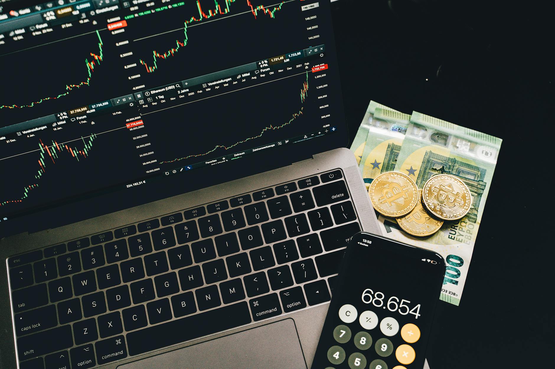 bitcoins and paper money beside a cellphone and laptop with graphs on screen